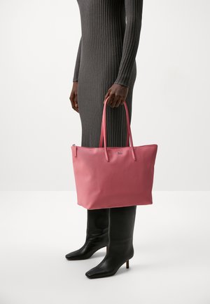 Sac fourre-tout rose texturé avec des poignées doubles et une fermeture éclair, tenu par une personne portant une robe grise moulante à côtes et des bottes noires jusqu'aux genoux.