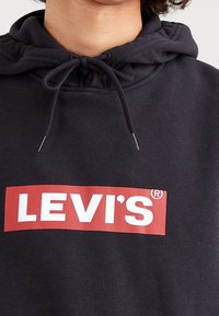 Černá mikina s kapucí, která má výrazné červené obdélníkové logo s nápisem "LEVI'S" bílými písmeny. Vyrobena z měkké látky.