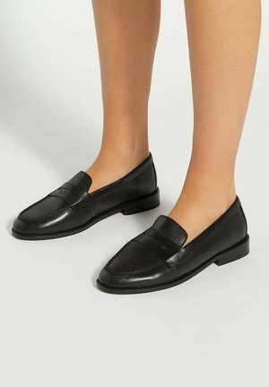 Personne portant des mocassins en cuir noir à enfiler avec un petit talon, debout sur un sol gris clair.