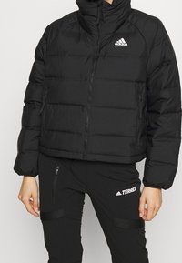 Giacca a vento nera con colletto alto, dotata di chiusure a zip e texture trapuntata. Il logo Adidas è visibile sulla parte superiore sinistra.