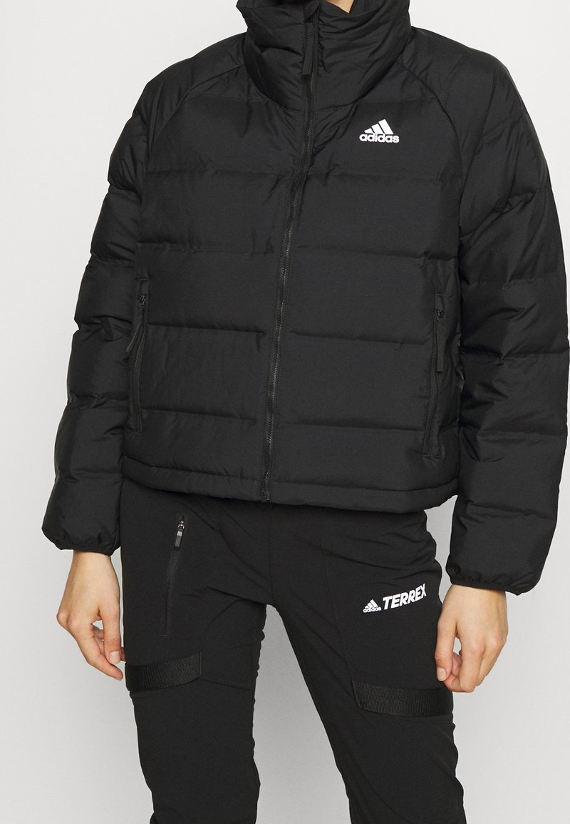 Giacca a vento nera con colletto alto, dotata di chiusure a zip e texture trapuntata. Il logo Adidas è visibile sulla parte superiore sinistra.