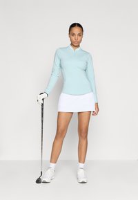 Camisola de golfe de manga longa azul claro com colarinho com fecho, combinada com uma saia branca. A modelo segura um taco de golfe e usa ténis brancos e luvas.