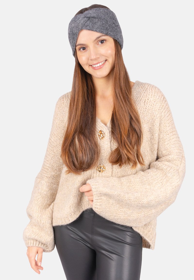 Cardigan en tricot chunky beige avec un col en V, trois boutons dorés et des manches surdimensionnées, associé à un pantalon noir ajusté et un bandeau gris.