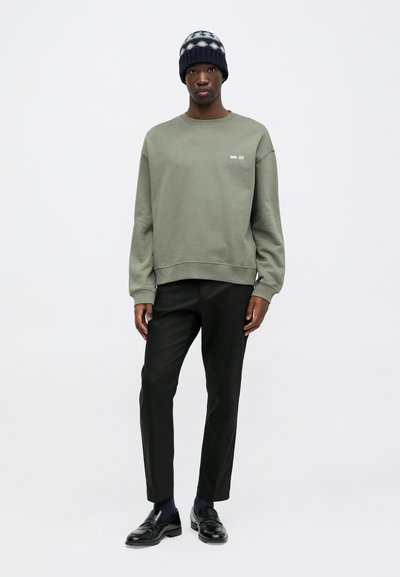 Olijfgroene sweatshirt met een ronde hals en geribde manchetten, gecombineerd met op maat gemaakte zwarte broeken en glanzende zwarte loafers. Marineblauwe gebreide beanie op het hoofd.