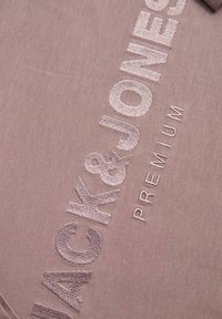Prim-plan cu material taupe cu text brodat ridicat „ACK&JONES PREMIUM” în fir asortat.