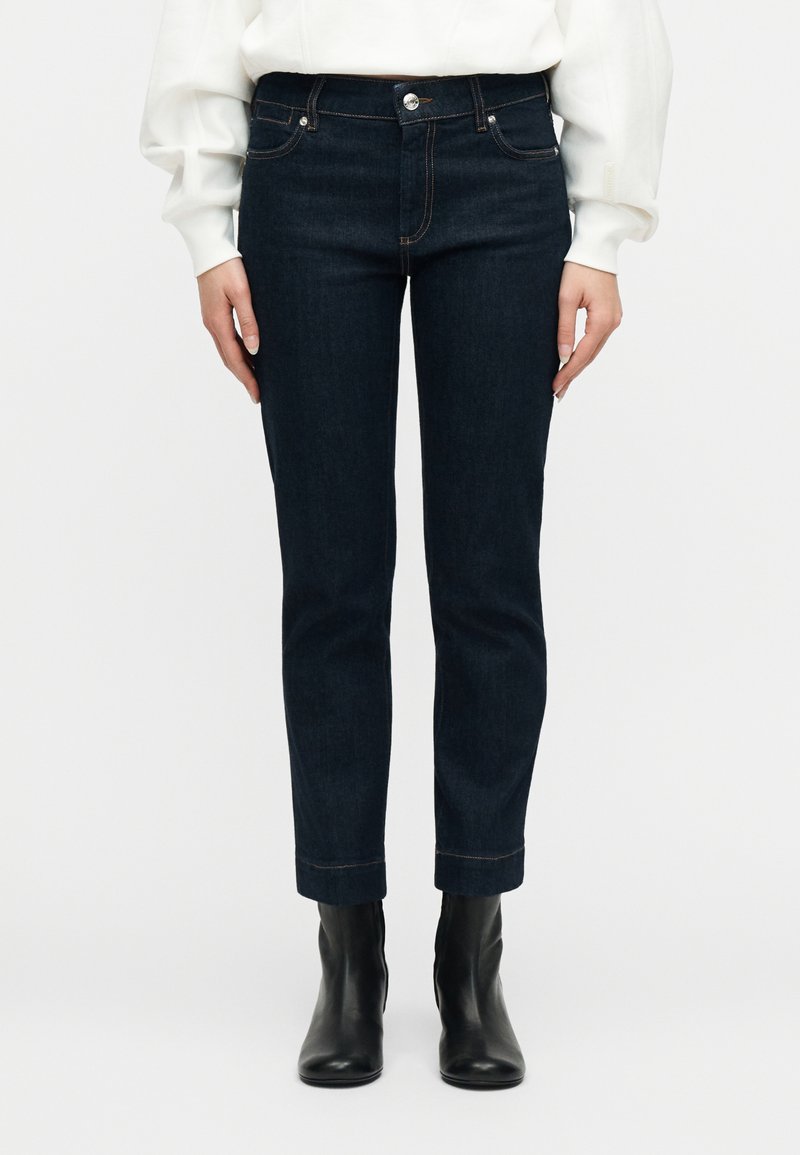 Γυναίκα που φοράει σκούρα μπλε τζι ν παντελόνια με ίσια γραμμή, λευκό μακρυμάνικο τοπ και μαύρα μποτάκια αστραγάλου, στέκεται μπροστά σε απλό φόντο.