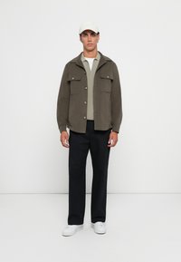 Olivgrüne Jacke mit zwei Brusttaschen, Knopfverschluss; hellgrauer strukturierter Pullover darunter; schwarze Hose; weiße Sneakers; heller Hut.