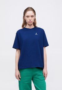 Granatowa bawełniana koszulka T-shirt z okrągłym dekoltem i krótkimi rękawami, z białym akcentem logo na piersi. Dopasowana do zielonych spodni.