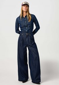 Denim-paitsi napituksella ja kahdella rinnataskulla yhdistettyinä leveälahkeisiin tummansiniset farkkuihin. Molemmissa on kontrastivärinen ompelu ja sileä rakenne.