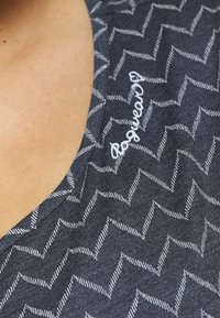 Námořnická modrá blůza s bílým zigzag vzorem, se zkoseným výstřihem a bíle vyšitým textem podél výstřihu. Měkká textura.
