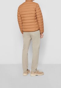 Giacca puffa tan con design trapuntato, pantaloni beige e scarpe di colore chiaro. La giacca presenta un colletto alto e una finitura leggermente lucida.