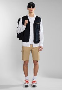 Gilet nero con diverse tasche sopra una camicia bianca, pantaloni cargo beige, cappellino nero, occhiali da sole e sneaker con accenti arancioni.