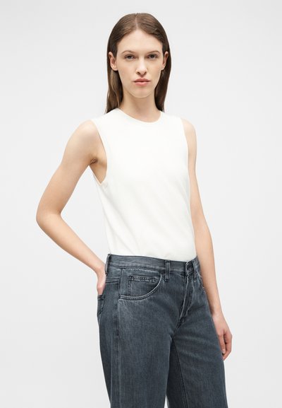 Jeune femme aux cheveux bruns lisses portant un haut blanc sans manches et un jean gris foncé taille haute, debout avec une main dans la poche.