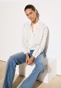 Weiße Bluse mit bestickten Details, langen Ärmeln und Knopfverschluss, kombiniert mit hellblauen Jeans. Einfaches Design, lässige Passform.