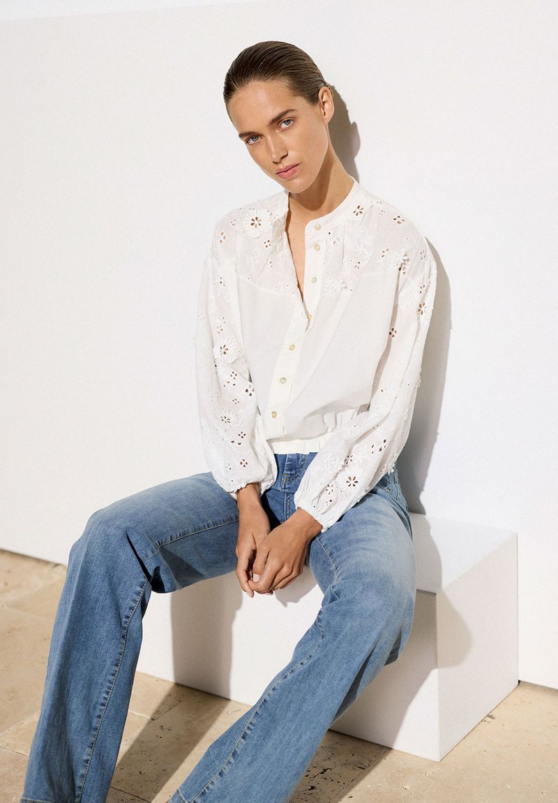 Weiße Bluse mit bestickten Details, langen Ärmeln und Knopfverschluss, kombiniert mit hellblauen Jeans. Einfaches Design, lässige Passform.