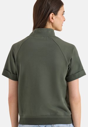 Frau mit langen dunklen Haaren, die ein olivgrünes kurzärmeliges Hemd mit Kragen trägt, von hinten vor einem schlichten Hintergrund gesehen.