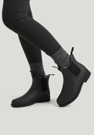 Ben iført mørkegrå leggings, grå sokker og sorte ankelregnstøvler med trækflapper på en lys grå baggrund.