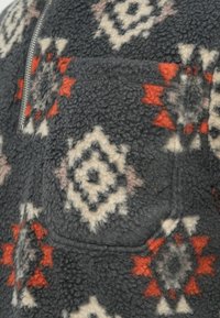 Fleece pulover tamno siva boja sa crvenim, bijelim i crnim geometrijskim uzorcima. Ima zatvarač na ovratniku i prednji džep. Mekana tekstura.