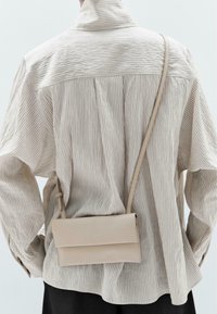 Borsa a tracolla beige con un design minimalista, forma strutturata, pelle liscia e patta pieghevole. Camicia a strisce testurizzata vista di spalle.