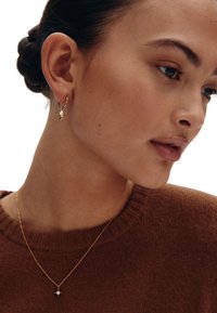 Goldene Creolen mit einem kleinen klaren Anhänger. Das Model trägt einen braunen Pullover mit Rundhalsausschnitt, der eine glatte Hautstruktur zeigt.