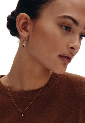 Guldörhängen med ring och en liten klar hängande detalj. Modellen bär en brun tröja med rund halsringning och visar en jämn hudstruktur.