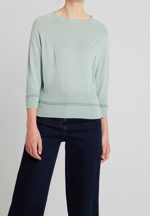 Femme portant un pull en maille bleu clair avec des motifs texturés et un jean large foncé sur un fond uni.