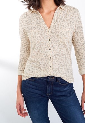 Femme portant une chemise crème boutonnée avec un petit imprimé floral et un jean bleu foncé, les mains détendues sur les côtés.