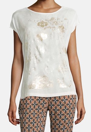 Femme portant un haut blanc à manches courtes avec un motif subtil en feuille d'or et un pantalon à motif géométrique en noir, blanc et marron.