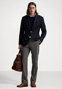 Granatowa marynarka z złotymi guzikami, szare spodnie, ciemny sweter i brązowe skórzane buty. Trzyma brązową skórzaną torbę. Szczegóły: okulary i długie włosy.