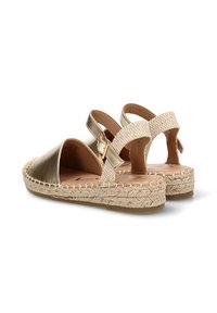 Par de sandalias doradas tipo espadrilles con suela de yute trenzado y correa beige para el tobillo con hebilla, mostradas desde la parte trasera sobre fondo blanco.