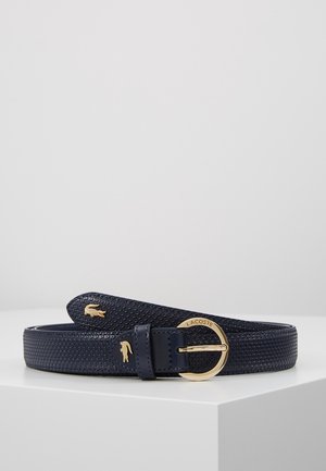 Ceinture en cuir texturé bleu marine avec logos crocodile Lacoste dorés et une boucle ronde dorée gravée « Lacoste », présentée sur une surface blanche.