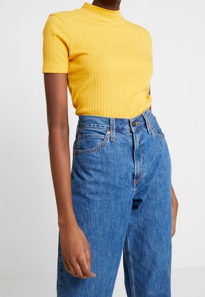 Osoba ubrana w żółty, prążkowany, krótki rękaw oraz niebieskie, wysokowalne jeansy, stojąca na tle jednolitego jasnego tła.