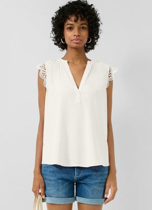 Kobieta ubrana w biały, bezrękawnik z koronkową lamówką i podwinięte dżinsowe szorty, trzymająca beżową torbę na jednolitym tle.