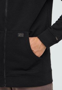 Sweat à capuche noir avec fermeture éclair, fabriqué en tissu doux, doté d'une poche avant, de poignets côtelés et d'une petite étiquette de marque sur le côté gauche.