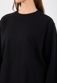 Čierny sweatshirt vyrobený z mäkkého bavlneného materiálu, s okrúhlym výstrihom a dlhými rukávmi so slučkovými manžetami. Hladká textúra a voľný strih.