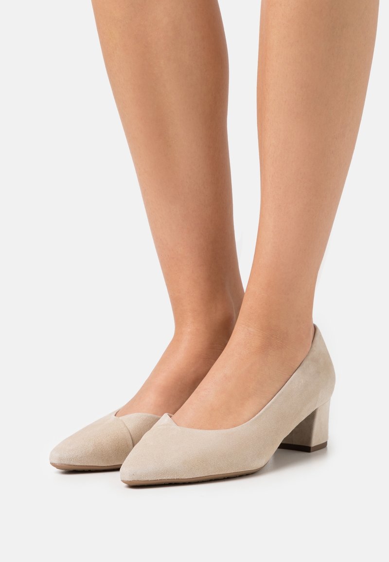 Escarpins en daim beige à talon bas carré portés pieds nus, bout pointu, texture lisse, fond neutre.