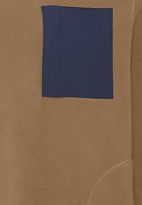 Sudadera de forro polar marrón con un bolsillo en el pecho de color azul marino. Presenta una textura suave y costuras sin costuras, resaltando el contraste en color y material.