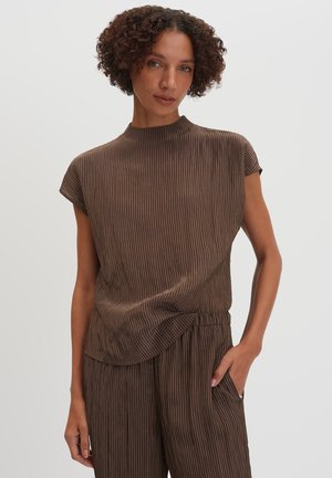 Vrouw met kort krullend haar, die een bruin gestreept topje met korte mouwen en bijpassende losse broek draagt, hand in de zak, neutrale achtergrond.