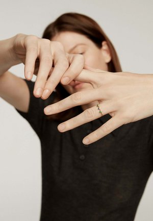 Una mujer con una camisa oscura estira los dedos de su mano izquierda, mostrando un delgado anillo de oro con piedras de colores en su dedo anular.