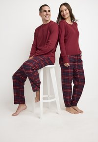 Camisas henley de manga larga rojas, combinadas con pantalones de pijama de cuadros en azul marino y rojo. La tela parece suave, con un corte holgado y tobillos ajustados.