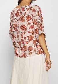 Kvinde iført en lys pink bluse med rustfarvet blomstermønster og en cremefarvet nederdel, vist fra ryggen mod en ensfarvet hvid baggrund.