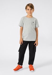 Jonge jongen die staat, gekleed in een grijs T-shirt met korte mouwen, zwarte cargobroek en feloranje sneakers tegen een effen achtergrond.