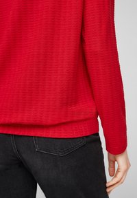 Red textured long-sleeve top with a wavy pattern, worn over black jeans. Visible hem and sleeve details.