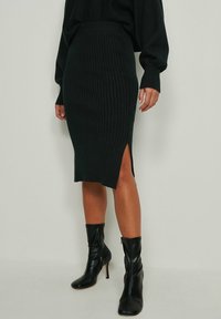 Nainen, jolla on päällään musta ribbiknitti midi-hame, jossa on sivuhaalari, sekä mustat korkokengät nilkkapituisina, seisoo yksinkertaista taustaa vasten.