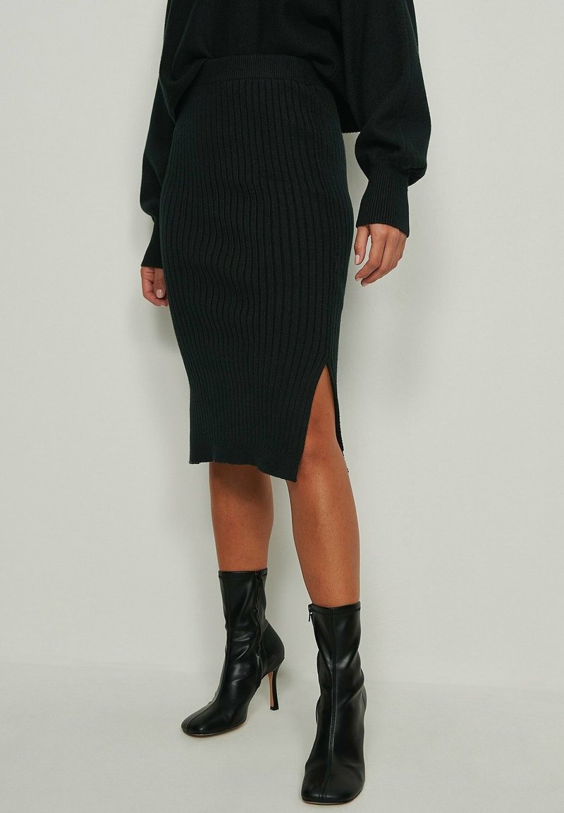 Nainen, jolla on päällään musta ribbiknitti midi-hame, jossa on sivuhaalari, sekä mustat korkokengät nilkkapituisina, seisoo yksinkertaista taustaa vasten.