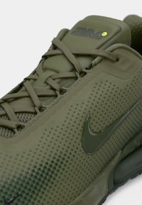 Gros plan d'une basket Nike vert olive avec une tige texturée, des lacets assortis et un logo Nike swoosh visible sur le côté.