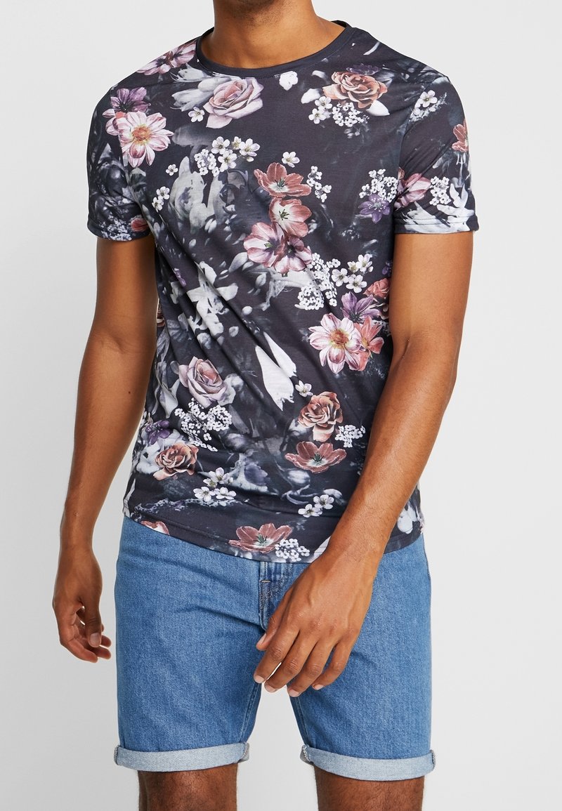 Homme portant un t-shirt à manches courtes à motif floral foncé et un short en jean bleu retroussé devant un fond uni.