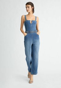Denim overal so širokými nohavicami, bez rukávov a s výstrihom v tvare kľúča. Obsahuje tkaný opasok a predné vrecká. Svetlomodrá farba.