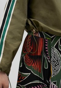 Olive green jacket with drawstring, featuring black and teal striped accents; paired with a pleated skirt with colorful floral patterns.