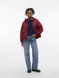 Kvinne med krøllete hår i en rød hettegenser, marineblå genser, blå jeans og svarte sko som står med en hånd i lommen.